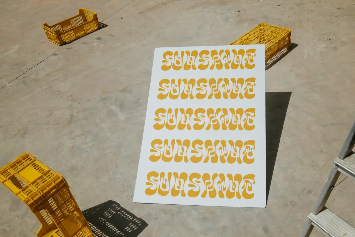Outdoor image of a large print of the word Sunshine repeated in a yellow wavy font, displayed on a board near yellow crates.