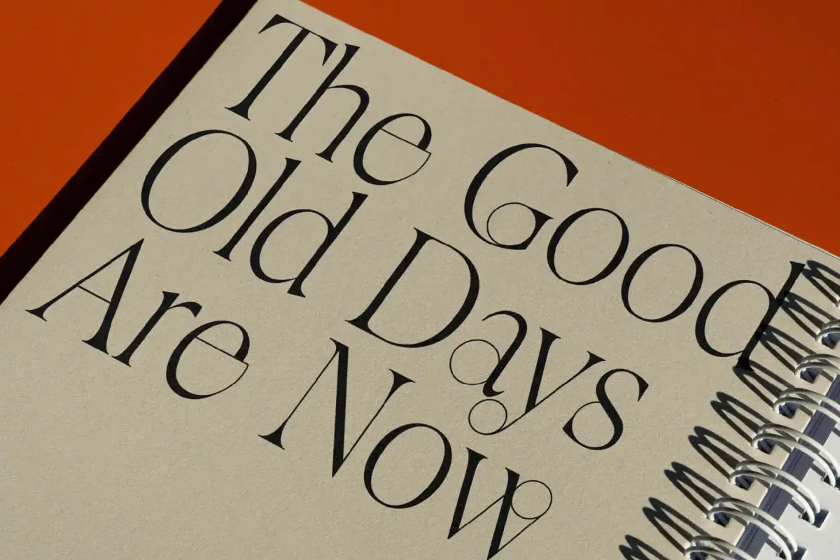 Close-up of a notebook cover with the phrase "The Good Old Days Are Now" in a sleek vintage serif font, set against a textured beige cover and paired with a spiral binding.