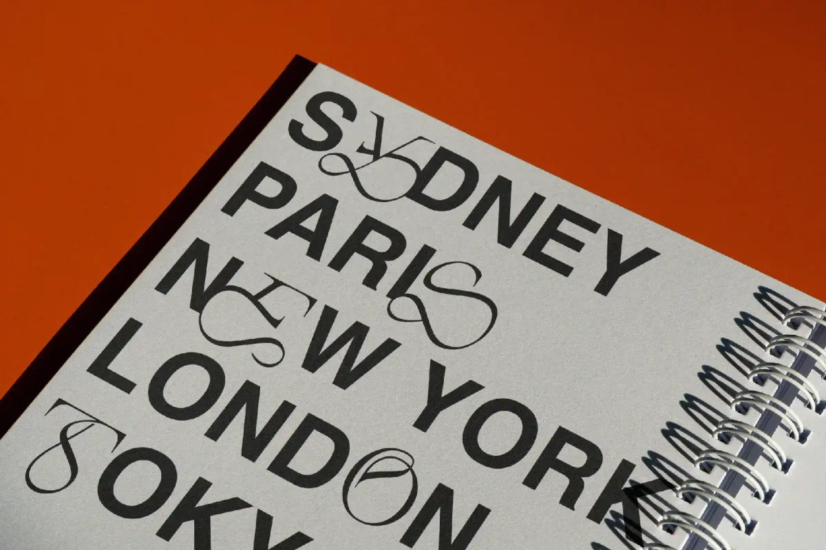 Cities like "Sydney," "Paris," and "New York" in bold serif fonts with decorative ligatures on a gray cover of a spiral-bound notebook.